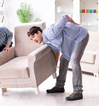 Two people trying to move a large chair with one of them experiencing back pain.