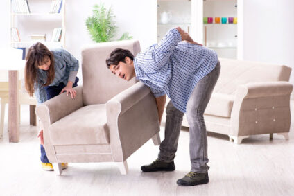 Two people trying to move a large chair with one of them experiencing back pain.