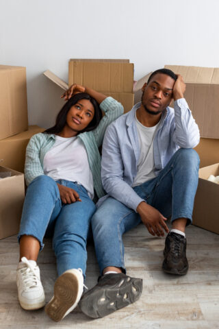 A frustrated couple sits on the floor surrounded by moving boxes.