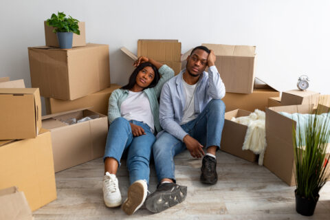 A frustrated couple sits on the floor surrounded by moving boxes.