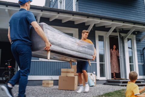 Two men carry a couch wrapped in plastic away from a house.