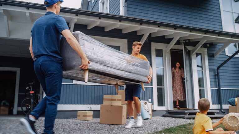 Two men carry a couch wrapped in plastic away from a house.