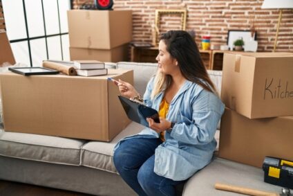 A woman sits on her couch holding a clipboard surrounded by moving boxes.