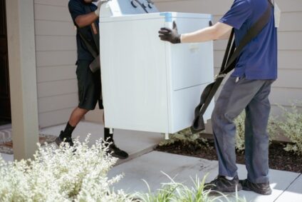 Two movers carry a washing machine out of a house.