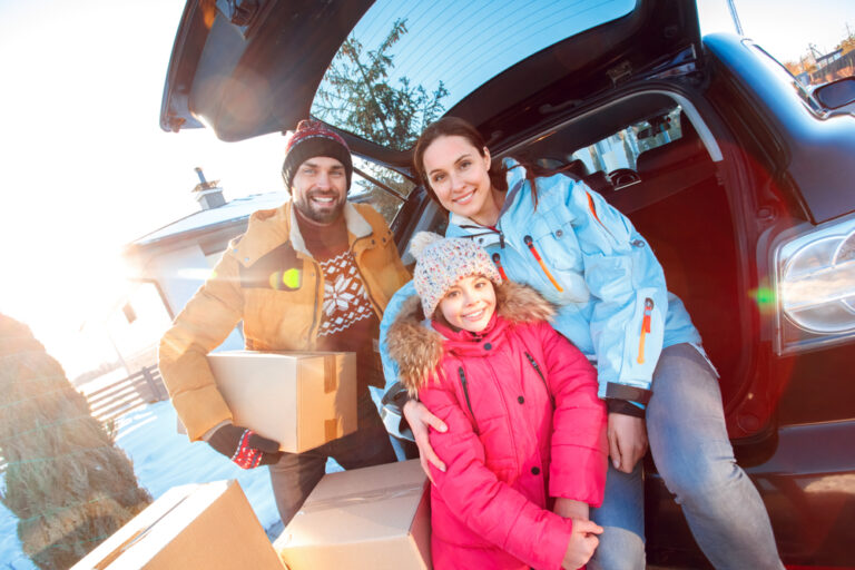 A young family smiles while wearing winter coats and packing moving boxes into their vehicle.