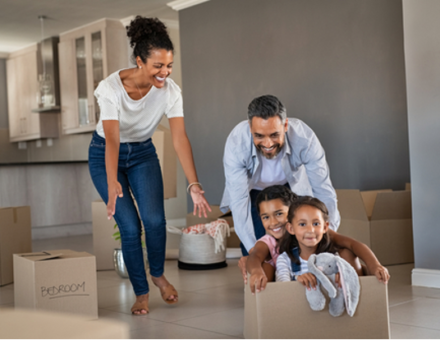 Smiling dad pushing excited daughters in cardboard box in new house while mom laughs