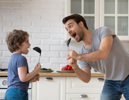 Parent and child sing in the kitchen using spoons as microphones.