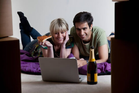 Young couple eats pizza on floor and watches movie on computer after moving in.