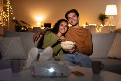 A couple cuddles on the couch with popcorn as they watch a movie.