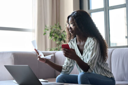 A woman sitting on the couch, holding a phone in one hand and a piece of paper in the other.