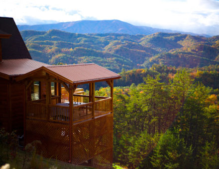 A log cabin sits high on a mountain overlooking a beautiful view of the forest below.