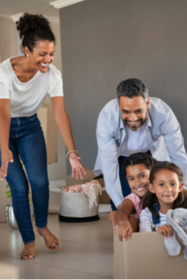 Smiling dad pushing excited daughters in cardboard box in new house while mom laughs