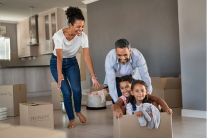 Smiling dad pushing excited daughters in cardboard box in new house while mom laughs