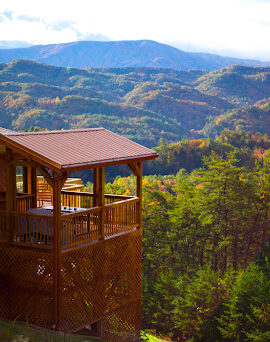 A log cabin sits high on a mountain overlooking a beautiful view of the forest below.