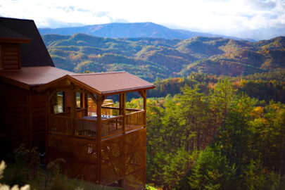 A log cabin sits high on a mountain overlooking a beautiful view of the forest below.