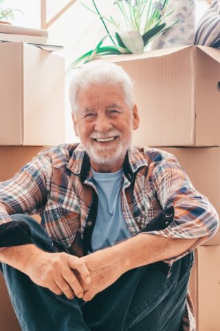 Happy senior man sits on floor near moving boxes.