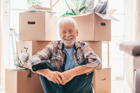 Happy senior man sits on floor near moving boxes.