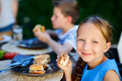 Kid smiling at the camera with a sandwich in her hand.