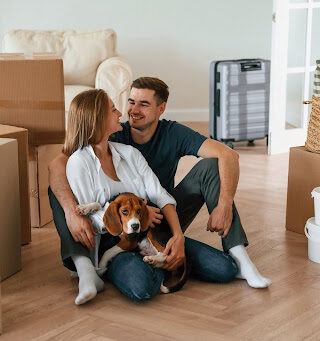 A couple with a dog surrounded by boxes and smiling.
