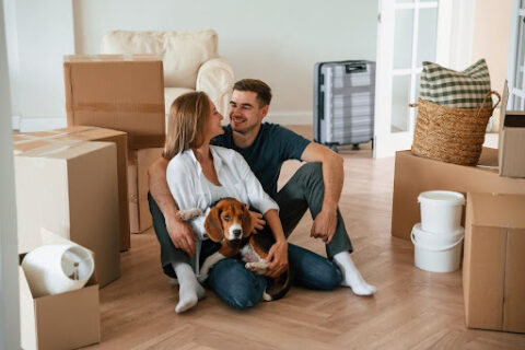 A couple with a dog surrounded by boxes and smiling.