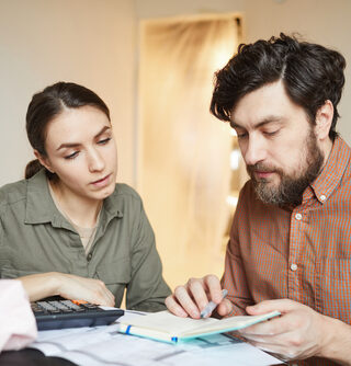 A couple looks down at a table with papers, a calculator, a notebook and a coffee on it. Their background is a white wall and open door.