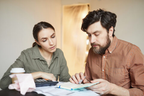 A couple looks down at a table with papers, a calculator, a notebook and a coffee on it. Their background is a white wall and open door.