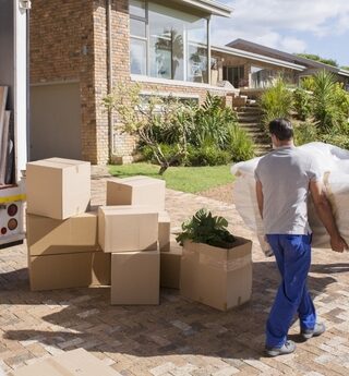 Two movers move a couch on a sunny day with a moving truck and boxes to the left.