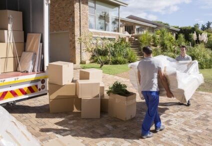 Two movers move a couch on a sunny day with a moving truck and boxes to the left.