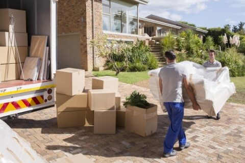 Two movers move a couch on a sunny day with a moving truck and boxes to the left.