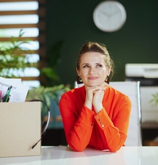 A woman wearing a bright orange shirt smiles while sitting at a desk with a box of office supplies.