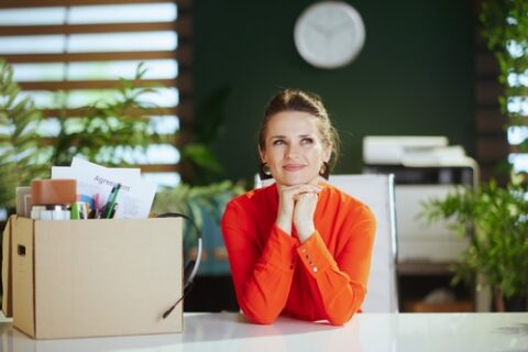 A woman wearing a bright orange shirt smiles while sitting at a desk with a box of office supplies.
