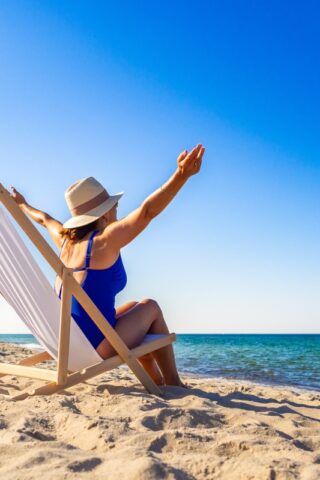 A woman sits on a beach chair in front of the ocean with her arms wide.